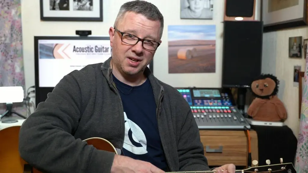 White middle aged man holding a guitar in his living room and talking to a camera